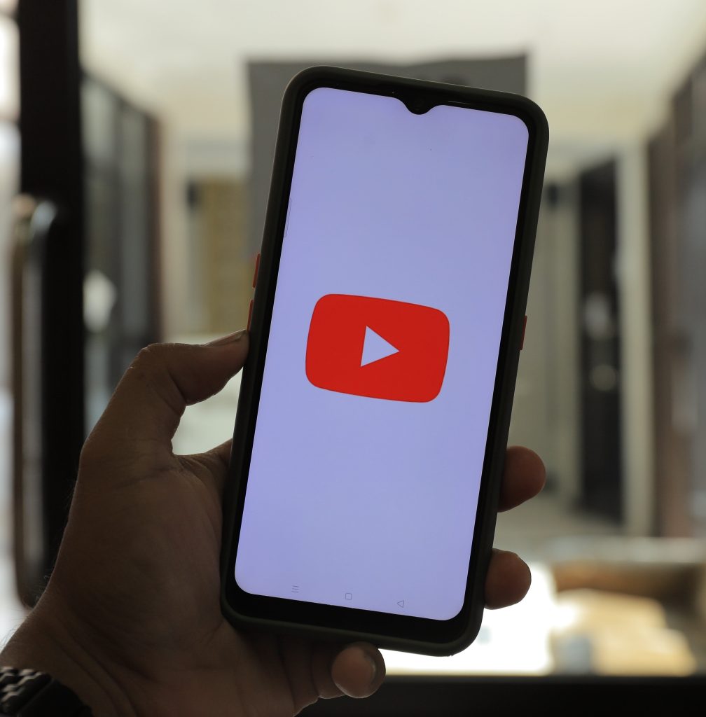Zu sehen ist ein Smartphone, welches mit dem Display zur Kamera gehalten wird. Auf dem Display des Smartphones ist das Youtube-Symbol auf weißem Hintergrund zu sehen.